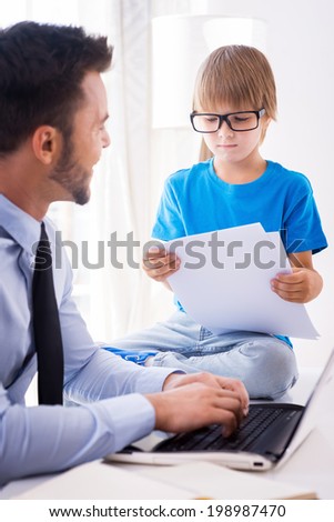 I want to be as smart as my father. Cheerful young man in shirt and tie working on laptop and looking at his son sitting close to him and examining documents