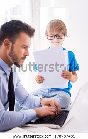 Busy working. Busy young man in shirt and tie working on laptop while his son in glasses sitting close to him and examining documents