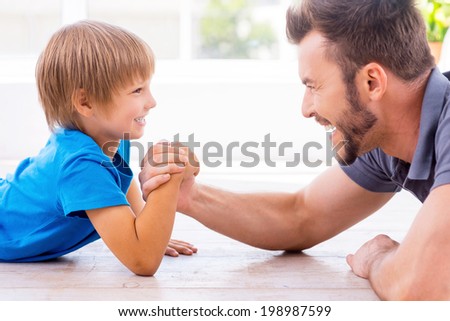Little champion in arm wrestling. Side view of happy father and son competing in arm wrestling while both lying on the hardwood floor