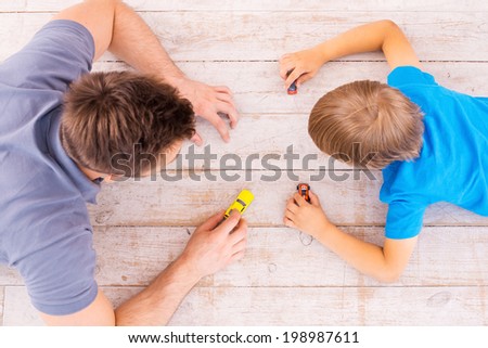 Playing together. Top view of father and son lying on the hardwood floor and playing with toy cars together