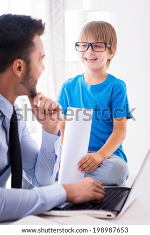 Working together is fun. Cheerful young man in shirt and tie working on laptop and looking at his smiling son sitting close to him and holding documents