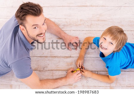 Playing together. Top view of happy father and son playing toy cars and looking up with smile while lying on the hardwood floor together