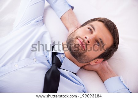 Lost in thoughts. Top view of handsome young man in shirt and tie holding hands behind head and smiling while lying in bed