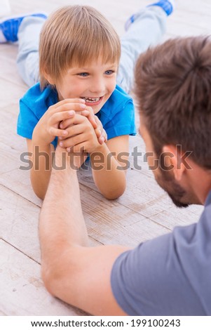I want to be as strong as my father! Top view of cheerful father and son competing in arm wrestling while both lying on the hardwood floor