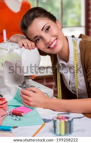 I love my job! Side view of beautiful female fashion designer leaning at the sewing machine and smiling while sitting at her working place