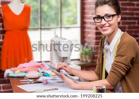 Fashion designer with digital tablet. Beautiful young woman working on digital tablet and smiling while sitting at her working place