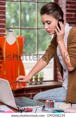 Busy fashion designer. Side view of confident young fashion designer pointing laptop with pencil and talking on the mobile phone while sitting at her working place