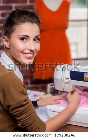 She is the best in fashion industry. Rear view of cheerful female fashion designer working on sewing machine and looking over shoulder while sitting at her working place