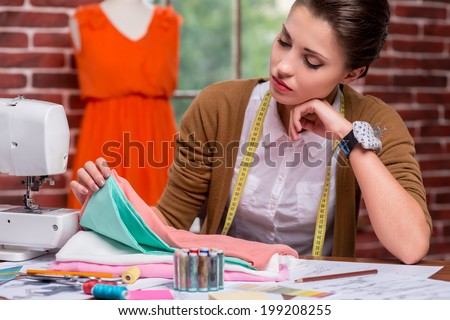 Examining textile before sewing. Thoughtful female fashion designer examining textile while sitting at her working place and with mannequin in the background