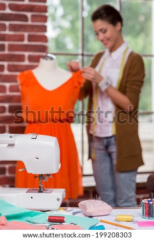 Enjoying creative work. Beautiful young fashion designer adjusting dress on the mannequin and smiling with sewing machine on the foreground