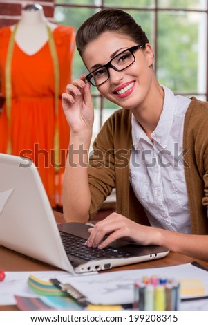 Beautiful fashion expert. Beautiful young fashion designer adjusting her glasses and smiling at camera while sitting at her working place