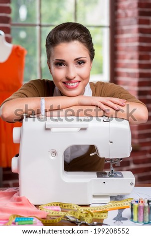 Confident fashion designer. Beautiful female fashion designer leaning at the sewing machine and smiling while sitting at her working place