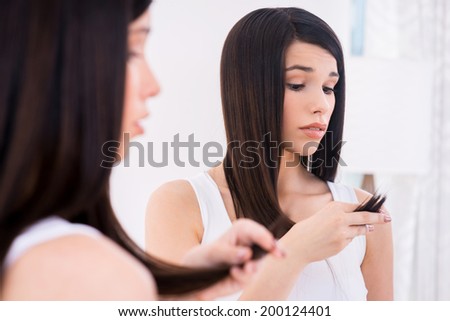 Examining her damaged hair. Frustrated young woman looking at her hair and expressing negativity while standing against mirror