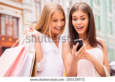 Can you imagine that? Two surprised young women holding shopping bags and looking at mobile phone together while standing outdoors
