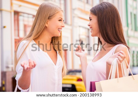 Friendly talk. Side view of two beautiful young women talking while carrying shopping bags and standing outdoors