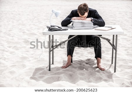 Tired of office work. Frustrated young businessman leaning his head at the table standing on sand