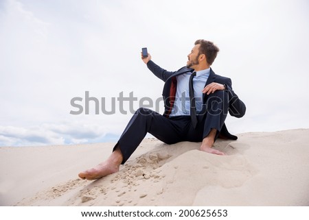 Poor signal. Frustrated young businessman searching for mobile phone signal while sitting on sand in desert