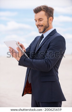 Surfing web in desert. Confident young man in formalwear working on digital tablet and smiling while standing in desert