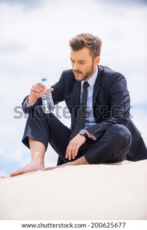 Water is life. Depressed young businessman holding bottle with water and looking at it while sitting on sand
