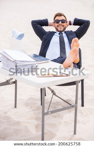 Time to relax. Cheerful young man in formalwear and sunglasses holding hands behind head and holding his feet on the table standing on sand