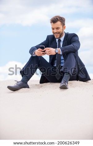 Staying in touch. Cheerful young businessman looking at his mobile phone and smiling while sitting on sand and against blue sky