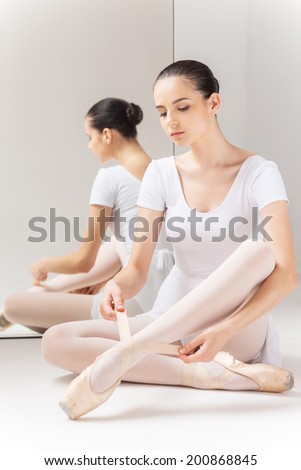 Preparing to show her best. Beautiful young ballerina in white tutu sitting against a mirror and tying her slippers