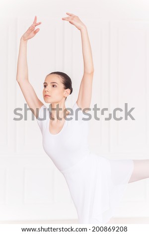 Beauty in her every move. Confident young ballerina in white tutu keeping arms raised while dancing in ballet studio