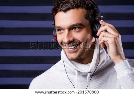 Music is my life. Handsome young man in white sweater adjusting his headphones and smiling while listening to the music and standing against striped background