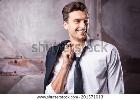 Relaxed handsome. Handsome young man in shirt and tie carrying his jacket on shoulder and looking away with smile while standing against metal background