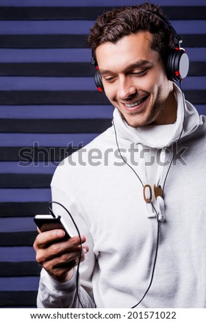 Choosing music to listen. Handsome young man in headphones holding MP3 Player and looking at it with smile while standing against striped background