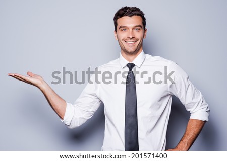Copy space on his hand. Handsome young man in shirt and tie holding copy space on his hand and smiling while standing against grey background