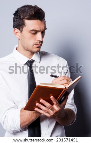 Writing down his ideas. Confident young man in shirt and tie writing something in note pad while standing against grey background