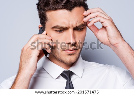 Bad news. Depressed young man in shirt and tie talking on the mobile phone and touching head with hand while standing against grey background