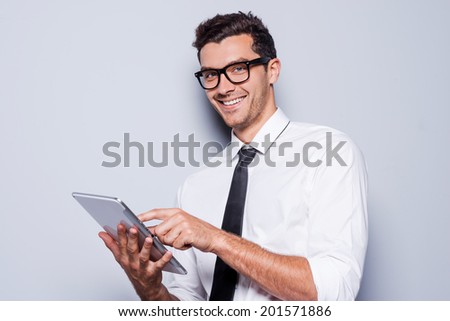 Businessman with digital tablet. Handsome young man in shirt and tie working on digital tablet and smiling while standing against grey background