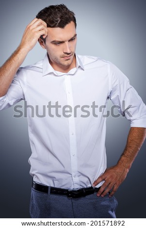 Lost in thoughts. Handsome young man in white shirt holding hand in hair and looking away while standing against grey background