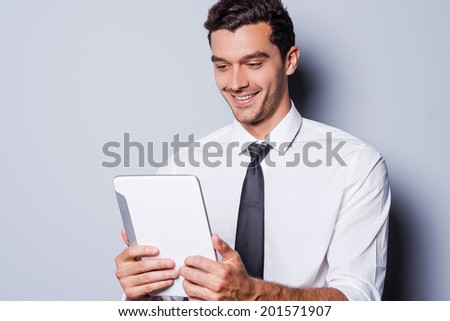 Good news! Cheerful young man in shirt and tie looking at digital tablet and smiling while standing against grey background