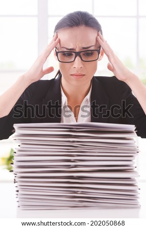 That is too much. Depressed young woman in suit tie looking at the stack of paperwork and holding head in hands while sitting at her working place