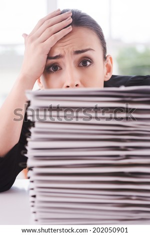 Tired of paperwork. Depressed young woman in suit looking out of the stack of documents laying on the table and touching her forehead with hand