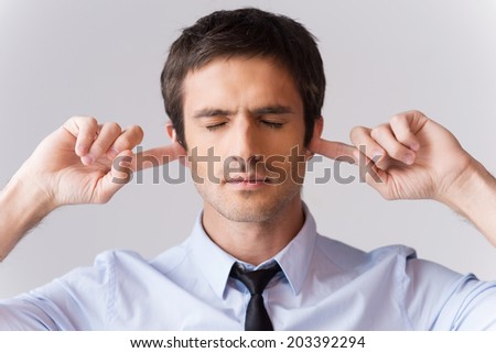 Too loud for me. Handsome young man in shirt and tie holding fingers in ears and keeping eyes closed while standing against grey background