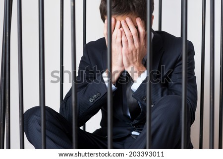 Business criminal. Depressed young man in formalwear standing behind a prison cell and looking at away