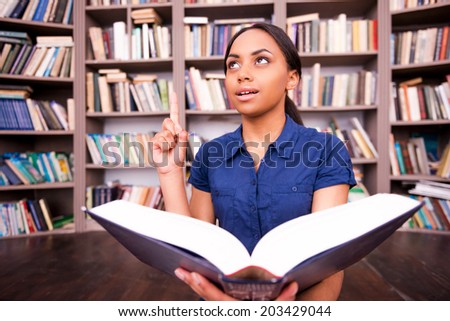 Just inspired. Surprised African female student holding a book and pointing up while sitting on the floor in library