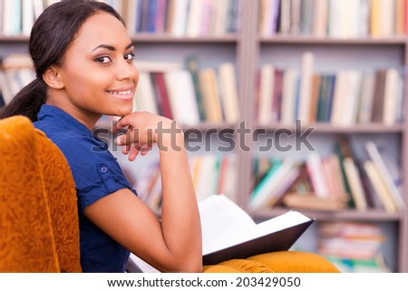 Reading a book in library. Rear view of beautiful African female student holding a book and looking over shoulder while sitting at the chair in library