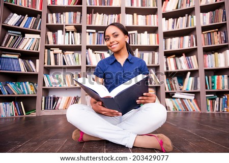 She loves reading. Beautiful African female student holding a book and smiling at camera while sitting on the floor in library