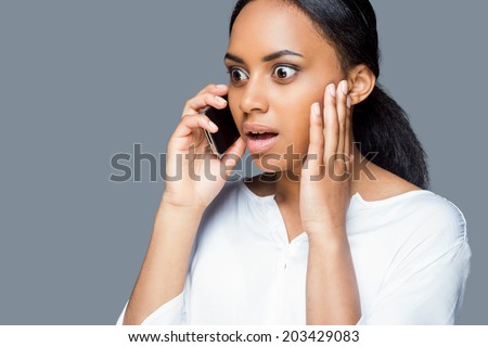 Unbelievable! Surprised young African woman talking on the mobile phone and keeping mouth open while standing against grey background