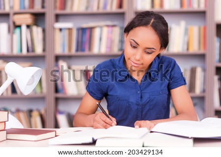 Student preparing to the exams. Confident young black woman writing something in her note pad and reading book while sitting at the library desk