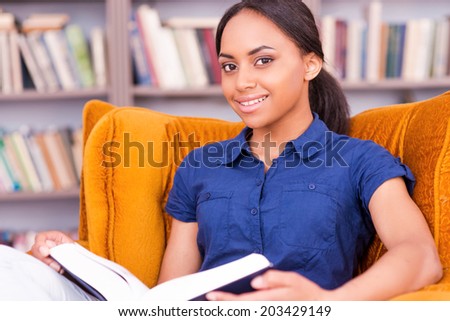 She found a peaceful place to read. Beautiful African female student reading a book and smiling while sitting at the chair in library
