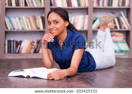 I love reading! Cheerful African female student reading a book while lying on the floor in library