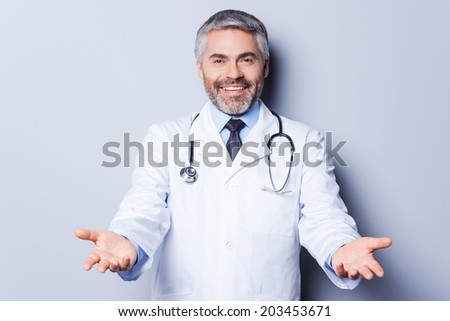Cheerful doctor. Cheerful mature grey hair doctor looking at camera while stretching out hands and smiling while standing against grey background