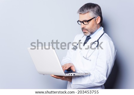Doctor with laptop. Confident mature doctor working on laptop while standing against grey background
