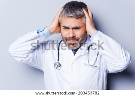 Stressed doctor. Frustrated mature doctor holding hands in hair and looking away while standing against grey background